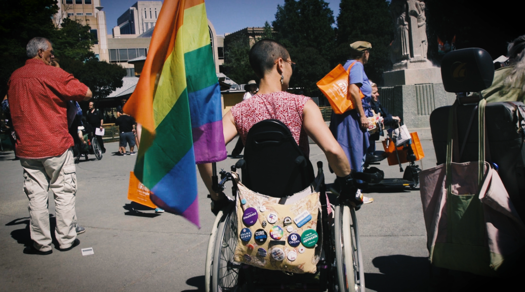 Image of the back of a person rolling away in a wheelchair with a bag on the back and a rainbow flag attached on the left side of the chair.