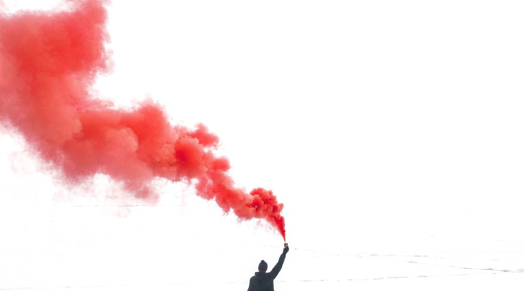 a silhouetted person stands on barren land holding a flare in the air. The image is black and white except for the flare, which is a string of cloudy red smoke drifting up and behind the person.