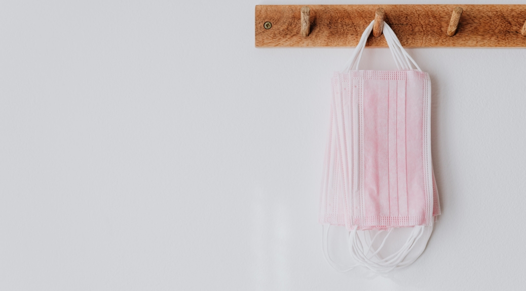 A picture of pink medical disposable face masks hanging on wood hooks on a white wall