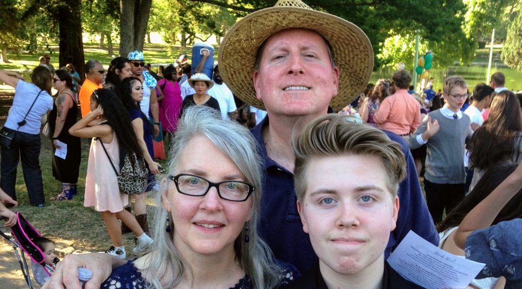 A family photo outside with a white mother, father, and son at his graduation from middle school. He wears a black robe. They all are smiling.
